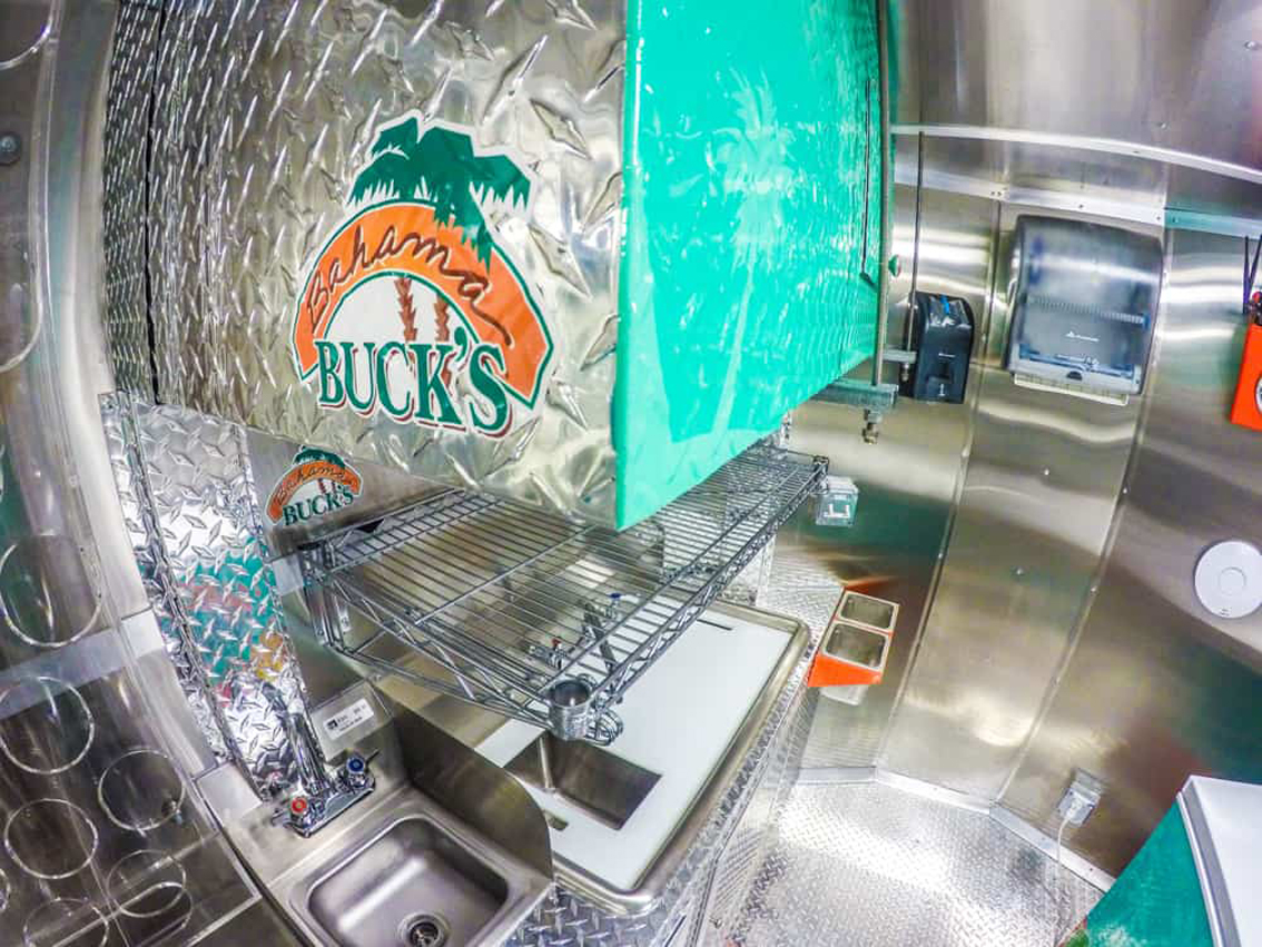 Bahama Bucks Shaved Ice Food Trailer by Cruising Kitchens is a custom-built trailer that provides high-quality shaved ice to customers. The sleek design and diamond plating make this trailer stand out, and the graphics package ensures that everyone knows where to go for a refreshing sno-cone.