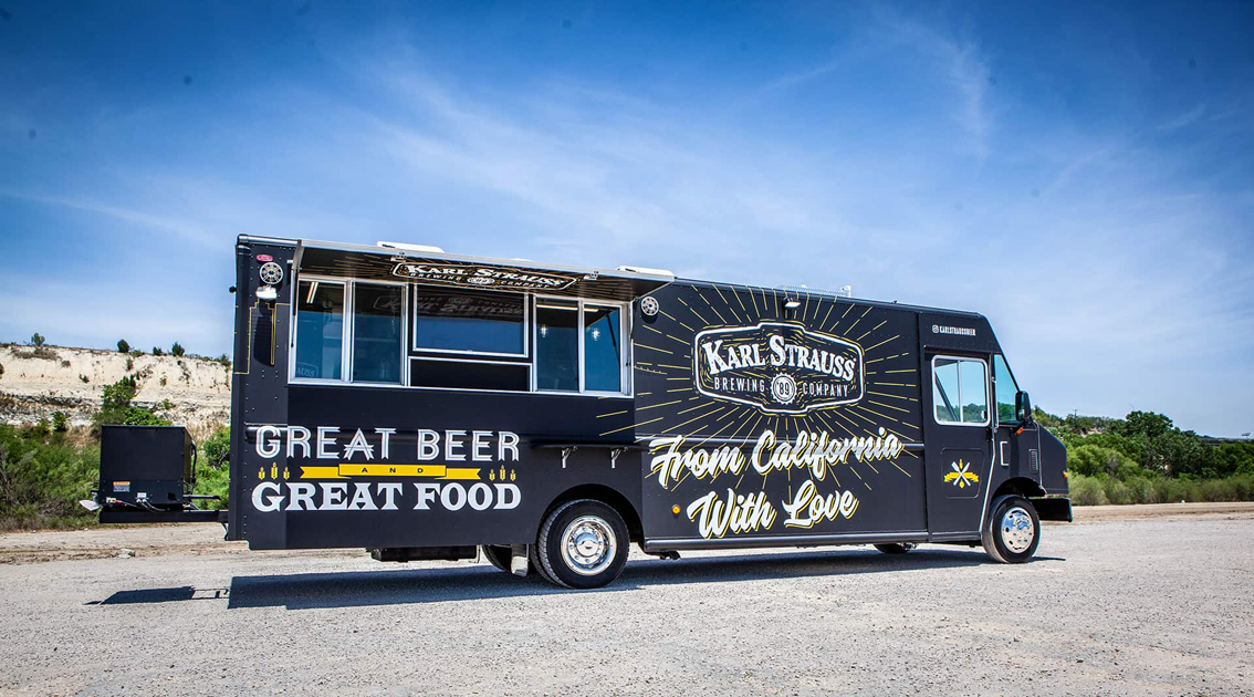 The Karl Strauss Food Truck is a unique mobile kitchen built by Cruising Kitchens, featuring the Karl Strauss logo with beautiful San Diego skyline backdrops. This top-of-the-line food truck comes equipped with commercial appliances for a quality culinary experience.